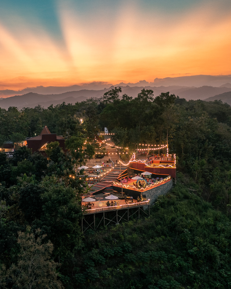 Puncak Saka: Näköalaravintola Indonesian Auringonlaskun Säteissä Ilmakuva Puncak Sakan rinneravintolasta tai -kahvilasta Indonesiassa, terassit valaistu valoketjuin, taustalla dramaattinen auringonlasku vuoristomaisemassa ja taivaalla näkyvät auringonsäteet.